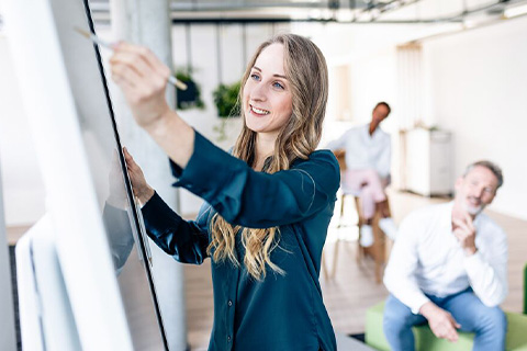 A woman in a meeting room with other people is working on a flipchart.