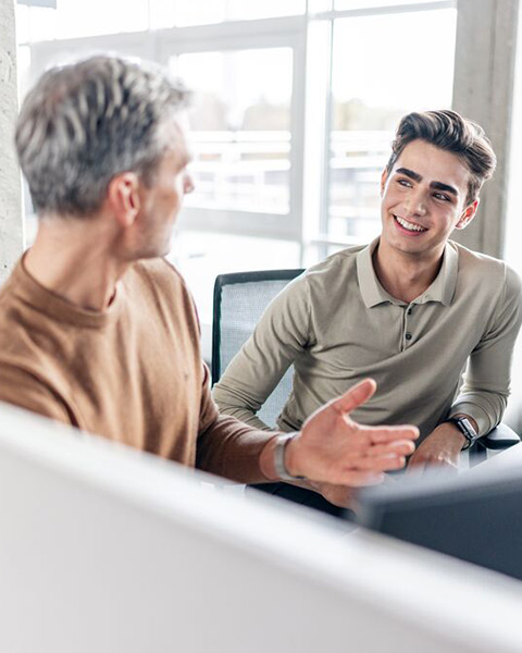 Ein älterer und ein junger Mann unterhalten sich sitzend vor einem PC.
