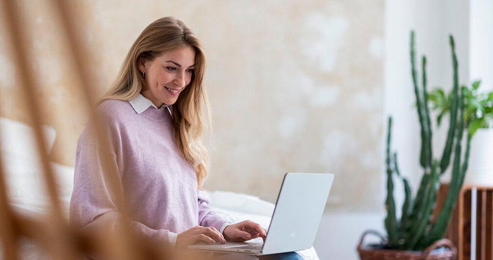 Eine junge Frau sitzt entspannt auf einem Bett und führt lächelnd ein Gespräch über ihren Laptop.