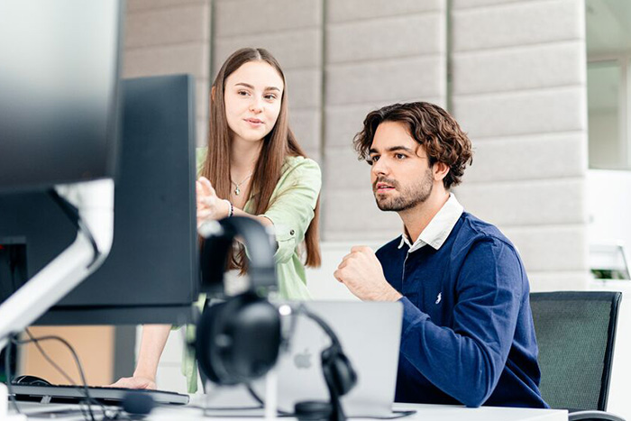 Zwei junge Menschen besprechen etwas und schauen dabei auf einen PC-Bildschirm.