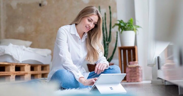 Eine junge Frau sitzt entspannt auf einem Bett und führt lächelnd ein Gespräch über ihren Laptop.