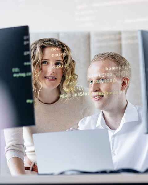 Zwei Personen schauen gemeinsam auf einen Monitor mit sichtbarem Code und arbeiten konzentriert an einem Laptop.
