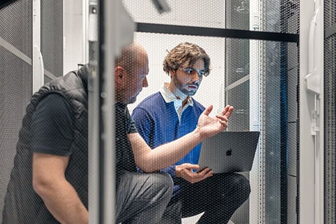 Zwei Männer diskutieren sitzend vor einem Server in einem Serverraum.